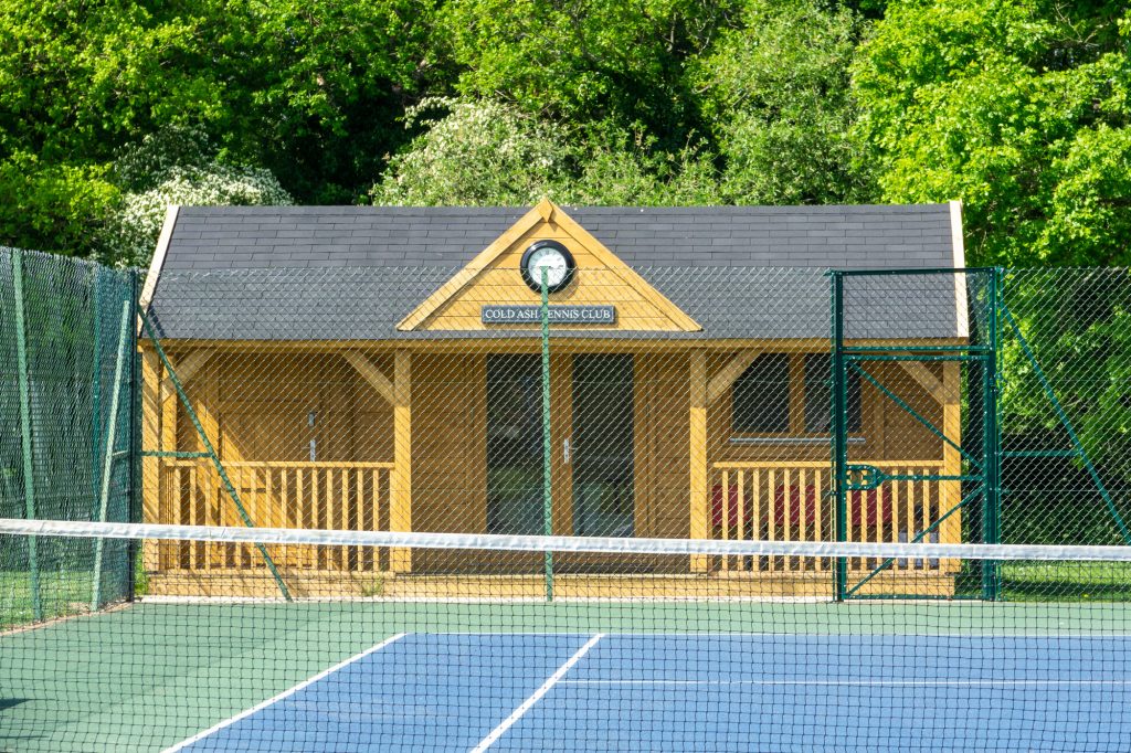 The tennis club pavilion is made of wood with grey roof tiles. A clock is visible on the front.